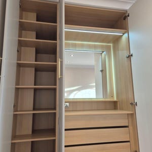 May include: A light wood-effect wardrobe with open doors, showing shelves, drawers, and a mirror. The wardrobe has multiple shelves on the left and a section with drawers and a mirror on the right. The interior is lit with a strip light.