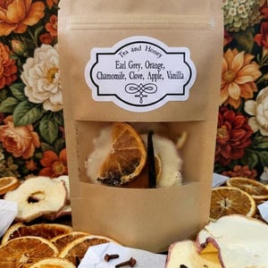 May include: A brown paper bag with a clear window, filled with dried orange slices, vanilla bean, and other ingredients. The bag has a label that reads "Tea and Honey" with a list of ingredients: Earl Grey, Orange, Chamomile, Clove, Apple, Vanilla.