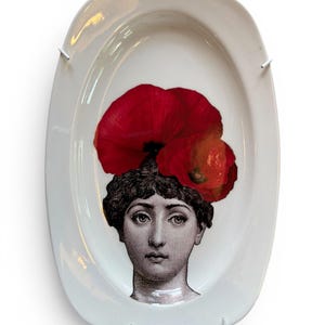 Op de afbeelding: Een ovale witte keramische schaal met een zwart-wit illustratie in gravurestijl van een hoofd en schouders. De figuur is versierd met twee grote, felrode klaprozen die direct boven op het donkere haar zijn geplaatst.