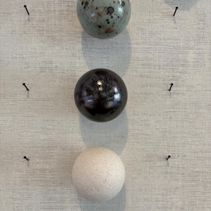Op de afbeelding: Drie bolvormige objecten verticaal gerangschikt. De bovenste bol is lichtblauw met donkerbruine spikkels. De middelste bol is glanzend donkerbruin metallic. De onderste bol heeft een getextureerd, mat gebroken wit oppervlak.