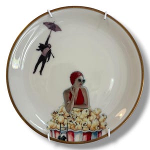 Op de afbeelding: Een wit keramieken bord met een goudkleurige rand, voorzien van een grafische illustratie van een persoon in een rood badpak en badmuts die uit een grote emmer popcorn komt, terwijl een figuur met een donkere paraplu linksboven zweeft.