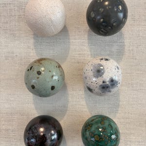 Puede incluir: Un conjunto de seis objetos esféricos de cerámica dispuestos en dos columnas. Las esferas presentan acabados variados que incluyen crema mate, carbón brillante con manchas claras, verde salvia moteado, blanco con marcas gris oscuro, marrón oscuro.