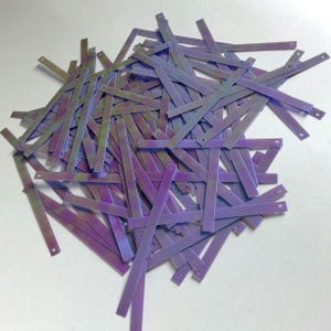 Könnte beinhalten: Ein Haufen aus schillernden lila und grünen rechteckigen Bastelstücken mit kleinen Löchern an den Enden. Die Teile sind verstreut und überlappen sich. Die Bastelmaterialien haben einen metallischen Glanz.
