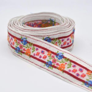 Pode incluir: Rolo de fita floral. A fita apresenta um fundo branco com uma borda vermelha e é decorada com flores azuis, laranja e rosa. A fita é provavelmente usada para artesanato, costura ou embrulho de presentes.