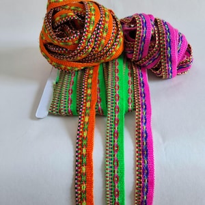 Op de afbeelding: Close-up van kleurrijke geweven linten in oranje, groen en roze. De linten zijn opgerold en afgerold, met ingewikkelde patronen en een verscheidenheid aan kleuren. De linten worden waarschijnlijk gebruikt voor knutselen of decoratieve doeleinden.