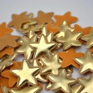 Può includere: Una collezione di abbellimenti a forma di stella in oro e arancione. Le stelle dorate hanno una lucentezza metallica, mentre le stelle arancioni hanno un aspetto strutturato, simile al feltro. Queste stelle decorative sono sparse.