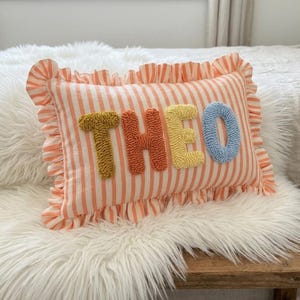 Könnte beinhalten: Ein rechteckiges Zierkissen mit pfirsichfarbenen und weißen Streifen und einem Rüschenrand. Der Name THEO ist in großen, strukturierten Punch-Needle-Buchstaben in Senfgelb, Orange, Gelb und Hellblau in der Mitte aufgestickt.