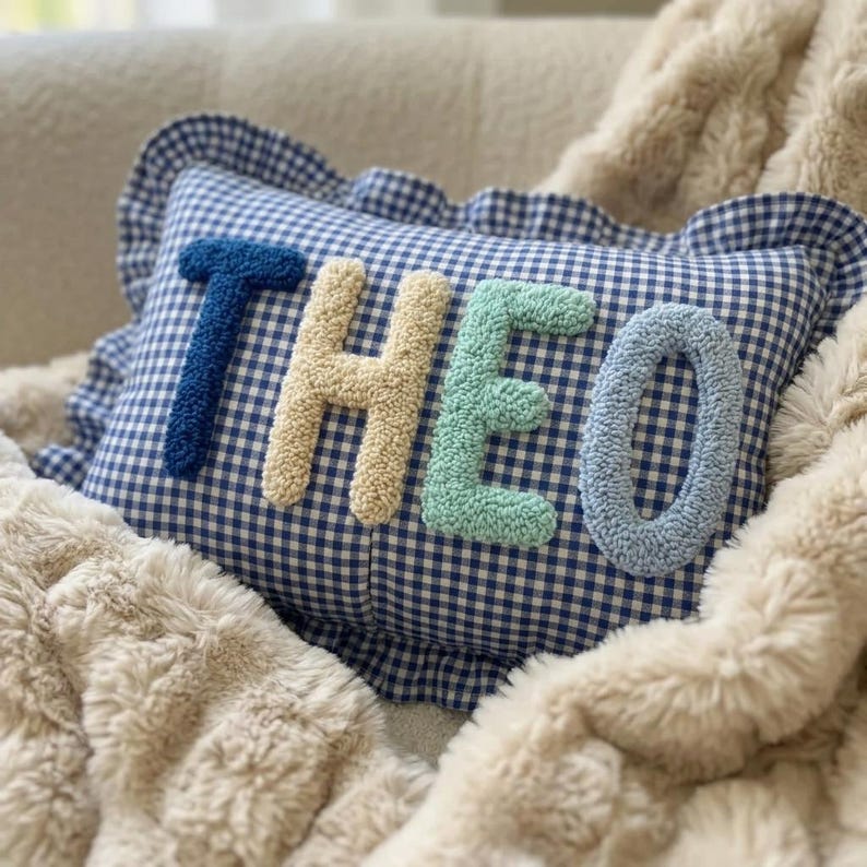May include: A blue and white gingham pillow with the name "THEO" embroidered in textured letters. The letters are in shades of blue, beige, and green. The pillow has a ruffled edge and is resting on a fluffy, cream-colored blanket.