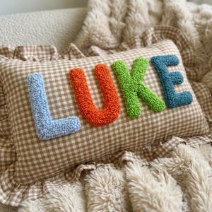 May include: A decorative pillow with the name "LUKE" embroidered in colorful, textured letters. The letters are blue, orange, green, and blue, set against a brown and white checkered background. The pillow has a ruffled edge.