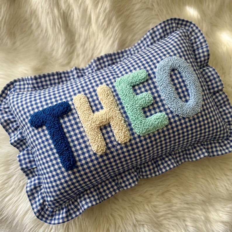May include: A blue and white gingham pillow with the name "THEO" embroidered in textured, colorful letters. The letters are in shades of blue, cream, and green. The pillow has a ruffled edge.