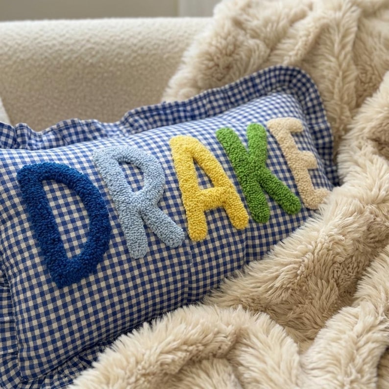 May include: A blue and white gingham pillow with the name "DRAKE" embroidered in colorful, textured letters. The pillow has a ruffled edge and is resting on a fluffy, cream-colored blanket.