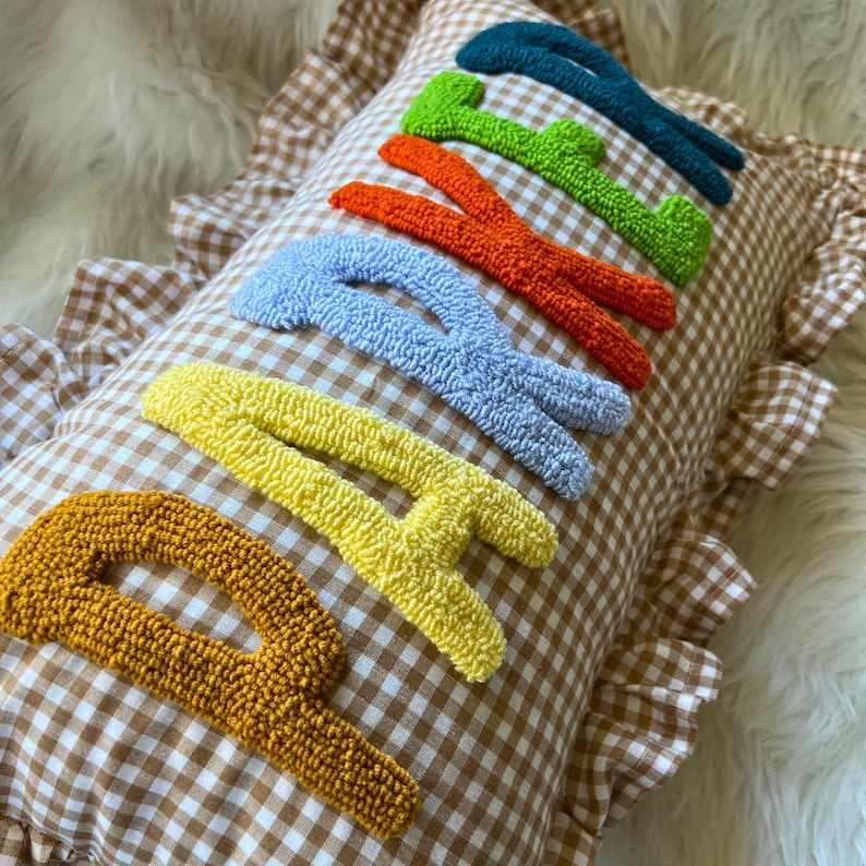 May include: A decorative pillow with the word "HAPPY" embroidered in colorful, textured yarn. The pillow has a brown and white checkered pattern and a ruffled edge.