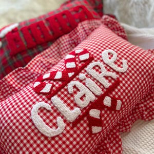 May include: Red and white gingham pillow with the name "Claire" embroidered in white. The pillow has a candy cane design and a ruffled edge. Another pillow with the name "Ellie" is visible in the background.