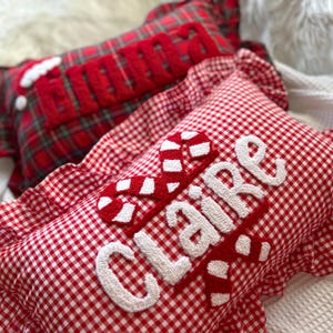 May include: Two decorative Christmas pillows. One is red and green plaid with the word "MERRY" in red. The other is red and white gingham with the name "CLAIRE" in white and red candy cane accents.