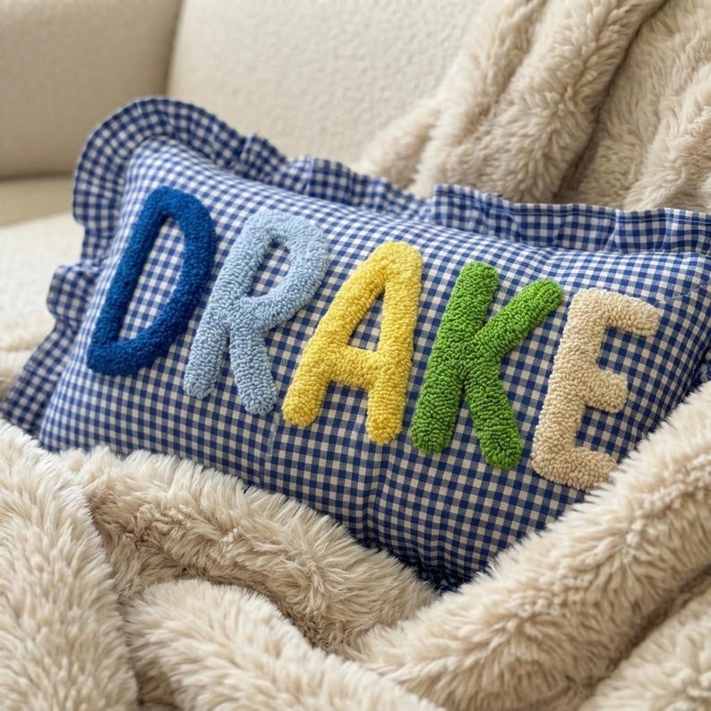 May include: A blue and white gingham pillow with the name "DRAKE" embroidered in colorful, textured letters. The pillow has a ruffled edge and is resting on a fluffy, cream-colored blanket.