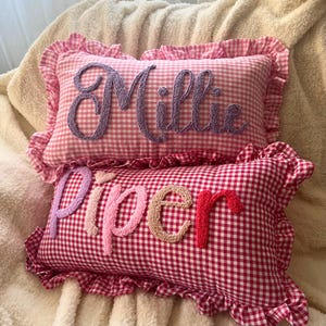 Könnte beinhalten: Zwei rechteckige Kissen mit Vichy-Mustern. Eines ist rosa und weiß, mit dem Namen "Millie" in Lila bestickt. Das andere ist rot und weiß, mit "Piper" in Rosa, Pfirsich und Rot bestickt.