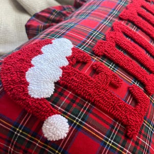 May include: A red and white Christmas pillow with a Santa hat design and the word "JOY" in red, textured letters. The pillow has a classic red plaid pattern with green and yellow accents, creating a festive holiday theme.