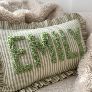 May include: A rectangular throw pillow featuring a light green and white vertical striped pattern. The name EMILY is embroidered in large, raised, sage green tufted letters across the centre. The pillow has a ruffled edge trim on all sides.
