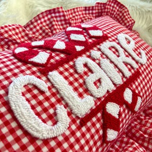 May include: A red and white gingham pillow with the name "CLAIRE" embroidered in white, accented with red and white candy cane designs. The pillow has a ruffled edge.