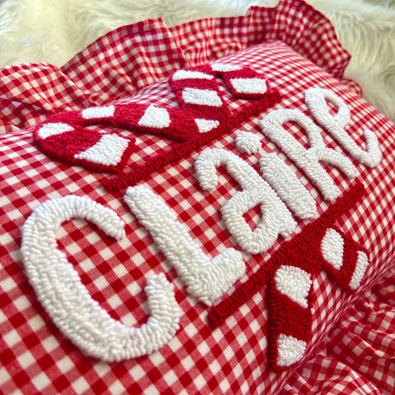 May include: A red and white gingham pillow with the name "CLAIRE" embroidered in white, accented with red and white candy cane designs. The pillow has a ruffled edge.