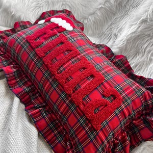 May include: A rectangular pillow with a red plaid pattern and a ruffled edge. The word "Believe" is embroidered in red, textured letters across the pillow. A small Santa hat is placed on the top.