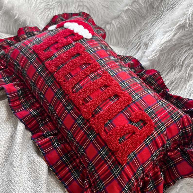 May include: A rectangular pillow with a red plaid pattern and a ruffled edge. The word "Believe" is embroidered in red, textured letters across the pillow. A small Santa hat is placed on the top.