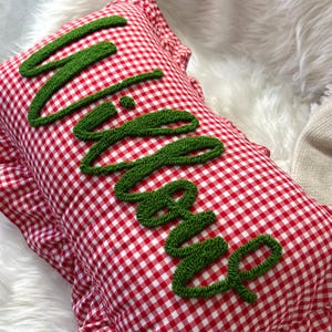 Könnte beinhalten: Ein rot-weiß kariertes Kissen mit dem Namen "Lila" in grünem, strukturiertem Garn gestickt. Das Kissen hat einen gerüschten Rand und liegt auf einer weißen, flauschigen Oberfläche.