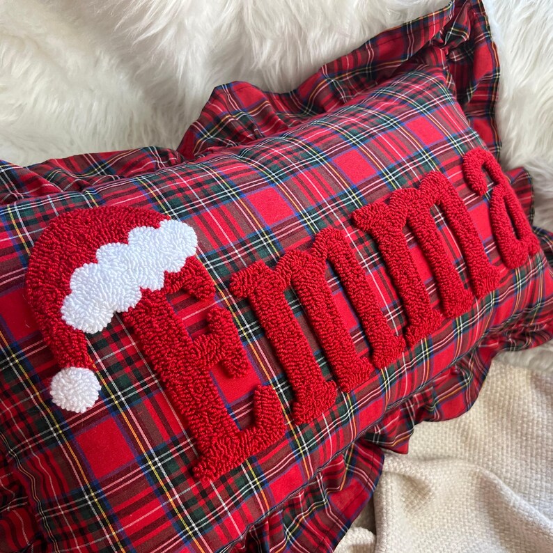 May include: A red plaid pillow with the name "Emma" embroidered in red, along with a Santa hat design. The pillow has a ruffled edge and is on a white fluffy surface.
