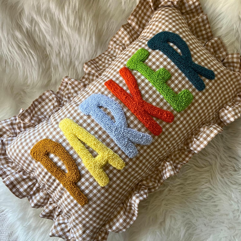 May include: A rectangular pillow with a brown and white gingham pattern and a ruffled edge. The word "PARKER" is embroidered in colorful, textured letters across the pillow's surface. The letters are in shades of yellow, blue, red, green, and teal.