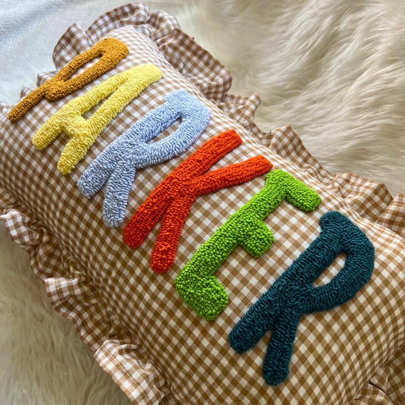 May include: A decorative pillow with the word "BAKER" embroidered in colorful, textured letters. The letters are in shades of orange, yellow, blue, red, green, and teal. The pillow has a brown and white checkered pattern and a ruffled edge.