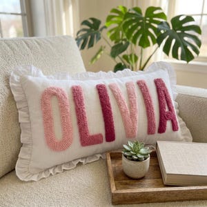 Könnte beinhalten: Ein rechteckiges weißes Zierkissen mit Rüschenrand, das den Namen OLIVIA in erhabener, texturierter Punch-Needle-Stickerei zeigt. Die Buchstaben wechseln sich in hellrosa, mittelrosa und dunkelmagentafarbenen Tönen auf dem weißen Stoff ab.