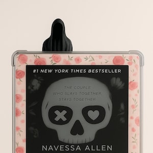 May include: An e-reader with a black cover featuring the book "Lights Out" by Navessa Allen. The cover has a skull graphic with an X and a heart for eyes. The e-reader is in a case with a floral pattern and a black hooded bookmark.