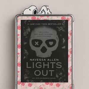 May include: An e-reader with a floral border and a Snoopy bookmark. The screen displays the book cover for "Lights Out" by Navessa Allen, a New York Times bestseller. The cover features a skull graphic.