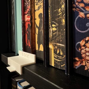 Puede incluir: Un sujetalibros blanco impreso en 3D con la palabra "BOOK" en el lateral, colocado en una estantería negra. El sujetalibros está junto a una fila de libros coloridos con lomos decorativos. Los libros son de varios tamaños y tienen portadas únicas.