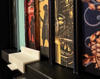 Könnte beinhalten: Ein weißer, 3D-gedruckter Buchhalter mit dem Wort „BOOK“ an der Seite, platziert auf einem schwarzen Bücherregal. Der Halter befindet sich neben einer Reihe farbenfroher Bücher mit dekorativen Buchrücken. Die Bücher sind unterschiedlich groß und haben einzigartige Cover.