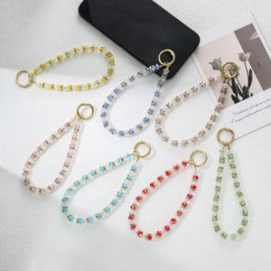 Puede incluir: Una colección de colgantes para teléfono con cuentas de varios colores, incluyendo amarillo, azul, rosa, turquesa, rojo y verde. Cada colgante tiene un anillo dorado para sujetarlo a un teléfono u otro accesorio.
