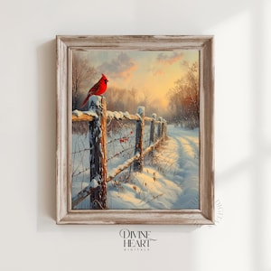 May include: A framed winter landscape painting featuring a vibrant red cardinal perched on a snow-covered wooden fence. The artwork depicts a snowy path leading into a forest, with a warm sunset in the background. The frame is a light, distressed wood.