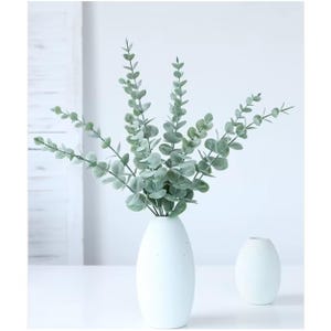 May include: A white ceramic vase holds a bundle of faux eucalyptus branches with round, silvery-green leaves. A smaller, matching vase sits beside it. The arrangement is set against a white background, creating a clean, minimalist aesthetic.