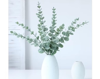 May include: A white ceramic vase holds a bundle of faux eucalyptus branches with round, silvery-green leaves. A smaller, matching vase sits beside it. The arrangement is set against a white background, creating a clean, minimalist aesthetic.