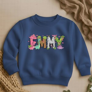May include: A navy blue sweatshirt featuring the name "EMMY" in a colorful, dinosaur-themed design. The letters are decorated with dinosaurs, flowers, and leaves. The sweatshirt is a crew neck style.