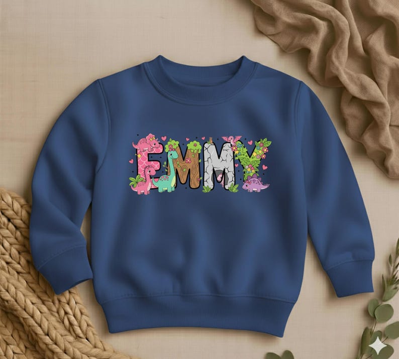 May include: A navy blue sweatshirt featuring the name "EMMY" in a colorful, dinosaur-themed design. The letters are decorated with dinosaurs, flowers, and leaves. The sweatshirt is a crew neck style.
