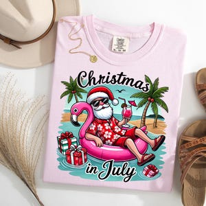 May include: A light pink t-shirt with the words "Christmas in July" and a graphic of Santa Claus relaxing on a flamingo float in a tropical setting. The shirt is next to a pair of brown sandals and a beige hat.