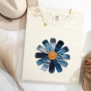 May include: A cream-colored short-sleeve t-shirt featuring a large daisy graphic made of various shades of blue denim fabric strips with a textured, circular, mustard-yellow center. The shirt has a Comfort Colors brand label at the inner neckline.