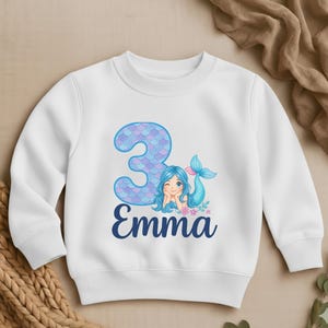 May include: White sweatshirt featuring a mermaid graphic with the number 3 and the name "Emma" in blue. The mermaid has blue hair and a pink tail. The number 3 is in a blue scale pattern.