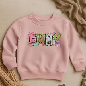 May include: A light pink sweatshirt with the name "EMMY" in large, colorful letters. The letters are decorated with cartoon dinosaurs, hearts, and floral accents. The sweatshirt has a crew neck and long sleeves.