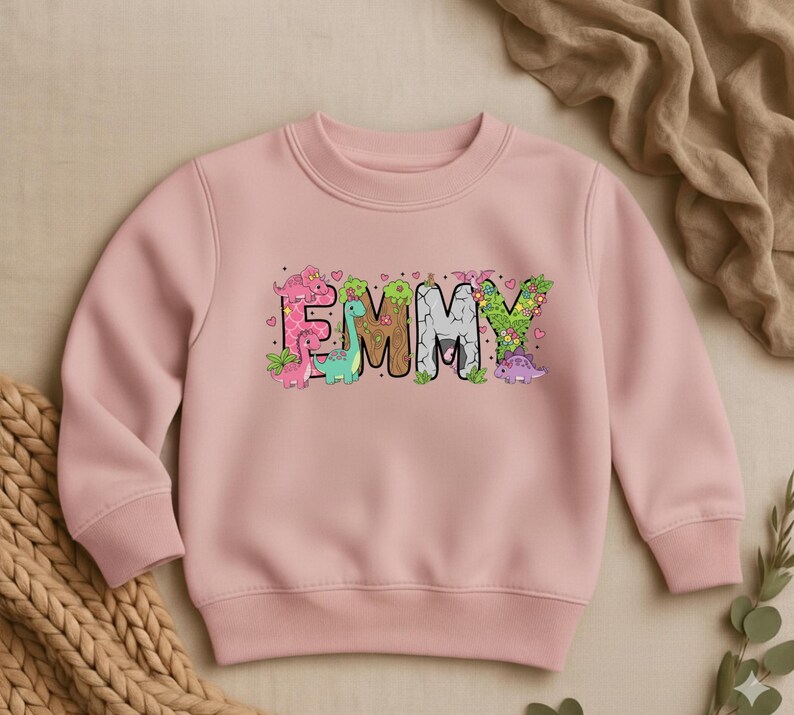 May include: A light pink sweatshirt with the name "EMMY" in large, colorful letters. The letters are decorated with cartoon dinosaurs, hearts, and floral accents. The sweatshirt has a crew neck and long sleeves.