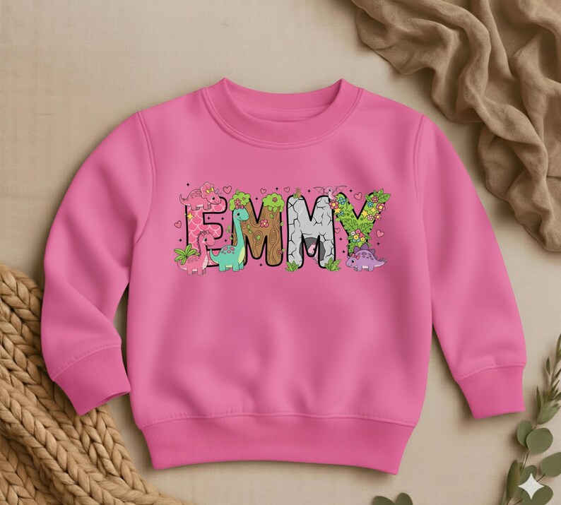May include: A pink sweatshirt with the name "EMMY" in colorful, dinosaur-themed letters. The letters are decorated with various dinosaurs and floral accents. The sweatshirt is made of a soft material and has a crew neck.