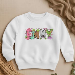 May include: White crewneck sweatshirt with the name "EMMY" in colorful, dinosaur-themed letters. The letters are decorated with pink, green, and brown dinosaurs, flowers, and hearts. A cute and playful design for kids.