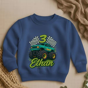 May include: A navy blue sweatshirt featuring a green monster truck with the number 3 and the name "Ethan" in a playful font. The design includes checkered flags and flames, perfect for a child's birthday.