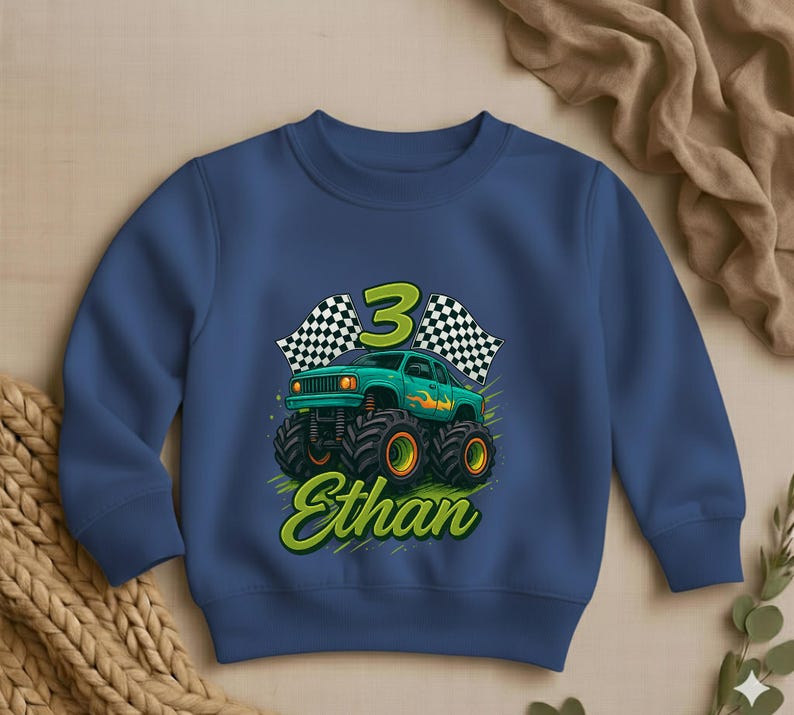 May include: A navy blue sweatshirt featuring a green monster truck with the number 3 and the name "Ethan" in a playful font. The design includes checkered flags and flames, perfect for a child's birthday.